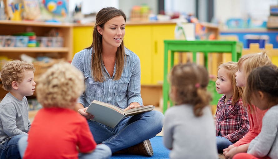A woman reads a book to a group of young children sitting in a circle in a classroom.