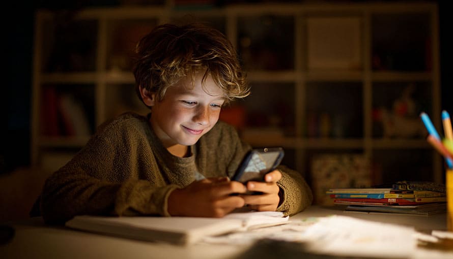A boy sits at a desk, smiling while looking at a smartphone, with books and papers nearby.